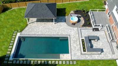 Aerial view of expansive paver patio with rectangular pool, hot tub, pergola structure, and surrounding manicured lawn.