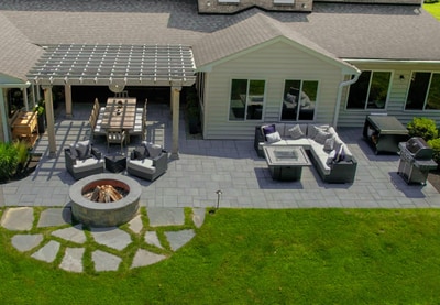 Multi-zone paver patio with pergola, fire pit, lounge seating, and landscape lighting integrated throughout, bordered by lawn.