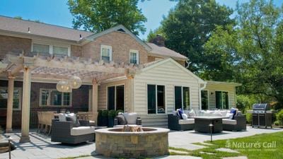 Stone fire pit anchors expansive paver patio with pergola, outdoor furniture, and modern home addition with foundation plantings.