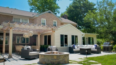 Stone fire pit anchors expansive paver patio with pergola, outdoor furniture, and modern home addition with foundation plantings.