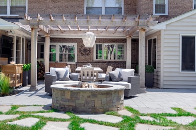 Flagstone patio with circular stone fire pit, wooden pergola overhead, lounge seating, and foundation plantings flanking brick home.