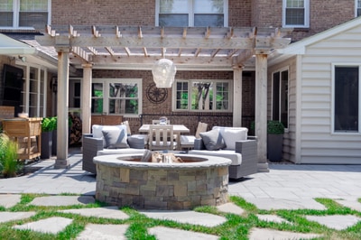Flagstone patio with circular stone fire pit, wooden pergola overhead, lounge seating, and foundation plantings flanking brick home.