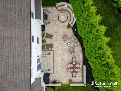 Aerial view of expansive paver patio with circular seating area, furniture arrangements, fire pit, and lush green hedging along perimeter.