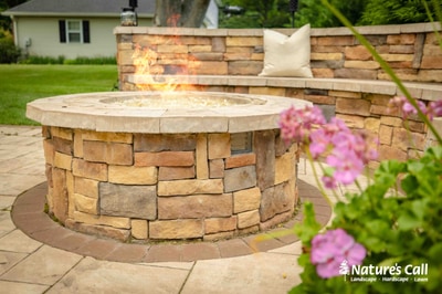 Round stone fire pit with cream-colored cap on paver patio, flanked by stacked stone seat wall and vibrant pink flowering plants.