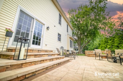 Large paver patio extending from house with stacked stone retaining wall, foundation plantings, and lounge seating at sunset.