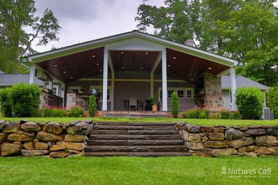 Brick pavilion with white columns and modern pitched roof, flanked by stacked stone retaining walls and manicured hedges lining the lawn.