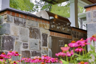 Outdoor kitchen featuring stacked stone counter, stainless steel grill and hood, with pergola overhead and bright pink flowering plants in foreground.