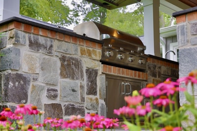 Outdoor kitchen featuring stacked stone counter, stainless steel grill and hood, with pergola overhead and bright pink flowering plants in foreground.