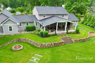 Modern home with covered porch, circular stone fire pit, curved stacked stone retaining wall, stepping stones, and lush green lawn landscaping.