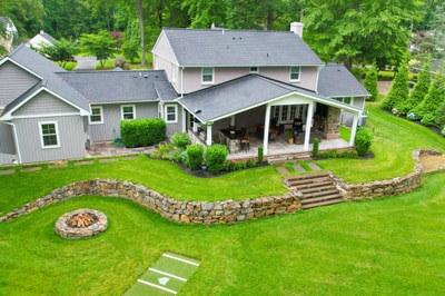 Modern home with covered porch, circular stone fire pit, curved stacked stone retaining wall, stepping stones, and lush green lawn landscaping.