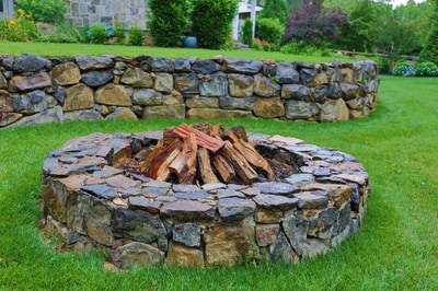 Stone fire pit with stacked fieldstone surround, burning wood interior, on lawn with residential stone wall in background.