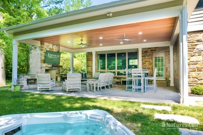 Covered patio with recessed lighting, stone columns, built-in fireplace, dining chairs, and dining table overlooking manicured lawn and hot tub.