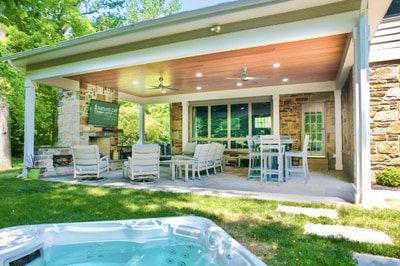 Covered patio with recessed lighting, stone columns, built-in fireplace, dining chairs, and dining table overlooking manicured lawn and hot tub.