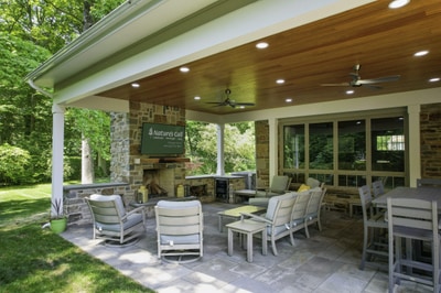 Expansive covered patio with recessed lighting, stone fireplace, stone columns, outdoor seating, and paver surface with wooded backdrop.
