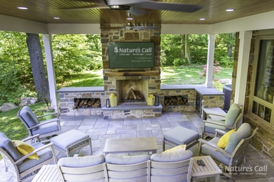 Covered patio with outdoor fireplace, stone construction, paver flooring, ceiling fan, recessed lighting, and comfortable seating surrounded by trees.