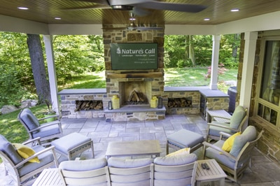 Covered patio with outdoor fireplace, stone construction, paver flooring, ceiling fan, recessed lighting, and comfortable seating surrounded by trees.