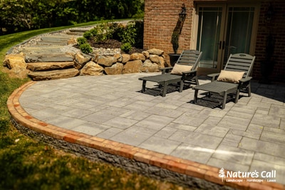 Curved paver patio with timber border, stone retaining wall with boulders, lounge chairs, and foundation plantings adjacent to brick home.