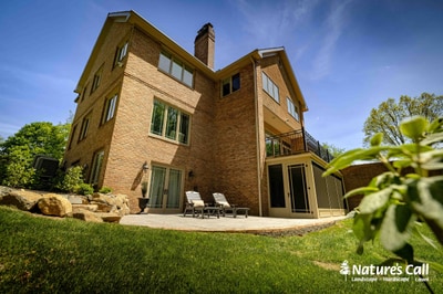Paver patio with large boulders at grade transition, lounge chairs, foundation plantings, and mature trees surrounding brick residential home.