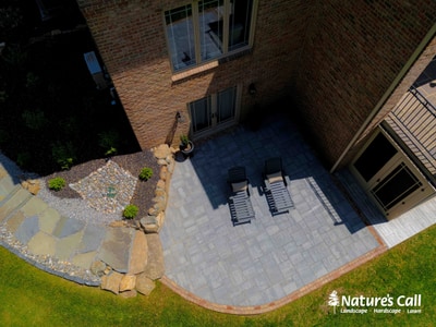 Curved paver patio with timber border, lounge seating, stone steps with boulder, specimen plant, and multi-level deck surrounding brick home.