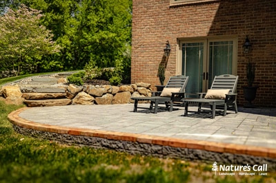 Paver patio with timber border, lounge chairs, stone retaining wall with boulders, foundation plantings, and shaded mature trees.