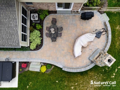 Overhead view of an expansive paver patio with curved edges, featuring a central dining table, lounge seating, and integrated planting beds with colorful shrubs.