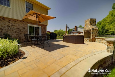 Tan paver patio with dining furniture and yellow umbrella, adjoining a stone fire pit surround and landscape plantings alongside a brick house.