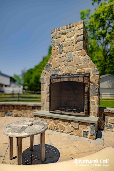 Stone outdoor fireplace with metal firebox surround, timber mantel, and decorative chimney cap, set on a tan paver pad with small side table.