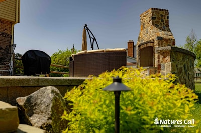 Outdoor entertaining space featuring a stone fireplace, dark metal grill surround, and bright green shrub plantings with landscape lighting in foreground.