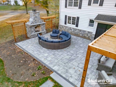 Gray paver patio with a circular fire pit surround featuring woven rattan construction and integrated seating, stone fireplace, and pergola.