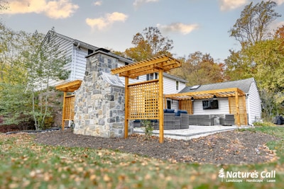 Stone and siding house with wooden lattice pergolas extending from the structure, leading to a modern outdoor kitchen with built-in appliances and dark countertop.