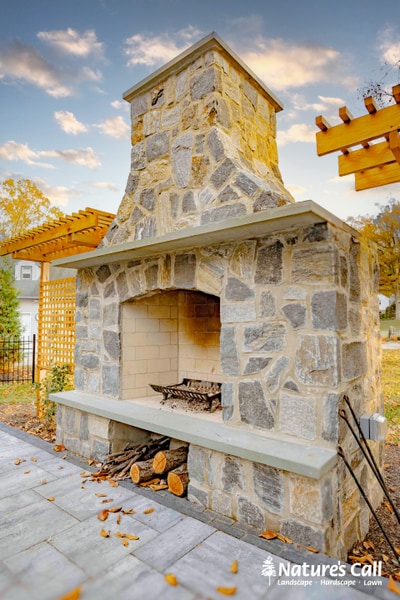 Tall stone fireplace with concrete mantel and tall chimney, situated on a paver patio with fireplace tools nearby and timber pergola visible to the side.