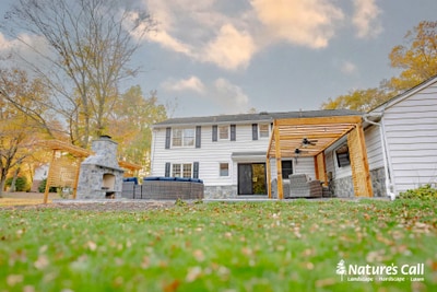 Wide landscape view of a white house with wooden pergola, stone planter beds, and paver patio, surrounded by mature trees and expansive lawn.