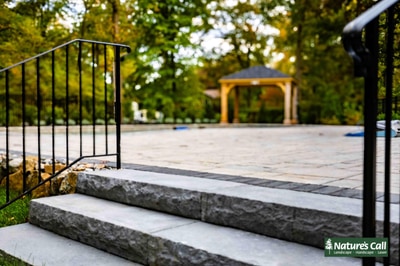 Bluestone steps with black metal railing lead to a paved patio featuring a wooden pergola structure, surrounded by mature landscape plantings.