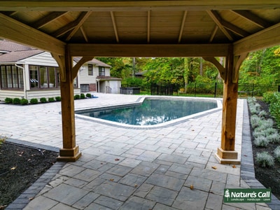 Wooden pergola overlooking paver patio with swimming pool, decorative plantings, and distant home with mature landscape backdrop.