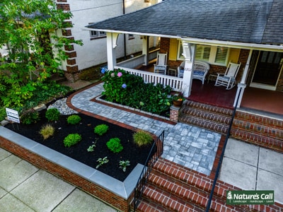 Curved paver walkway with brick planter beds and foundation plantings lead to brick-stepped entry with white cottage exterior.