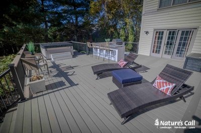 Composite deck with metal railings, outdoor kitchen, lounge seating, and accent planters overlooks mature wooded landscape.