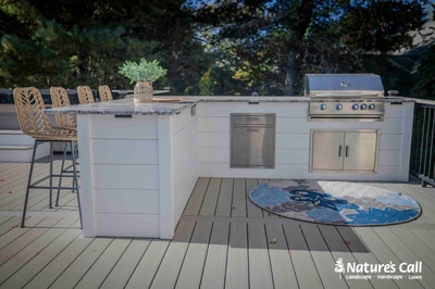 Built-in outdoor kitchen with stainless steel grill and appliances, bar seating, and decorative table on elevated composite deck.