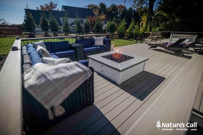 Composite deck with black metal railings features fire pit, sectional seating with blue cushions, and mature shade trees framing home.