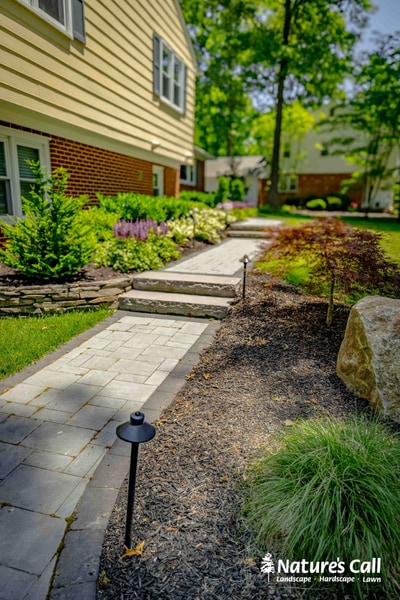 Staggered paver stepping stone walkway with landscape lighting, mulch beds, and ornamental grasses flanking brick home foundation.