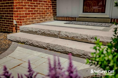Natural stone steps with timber risers leading to paver patio against brick home, accented by flowering shrubs.