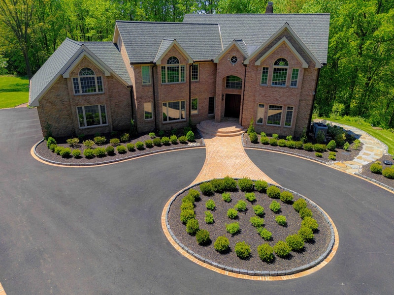 Eye-level view of the Hockessin home's front facade: the multi-gable brick estate with arched windows sits centered behind the curved paver driveway, with the boxwood-planted oval and front beds in the foreground and a flagstone path visible at the right