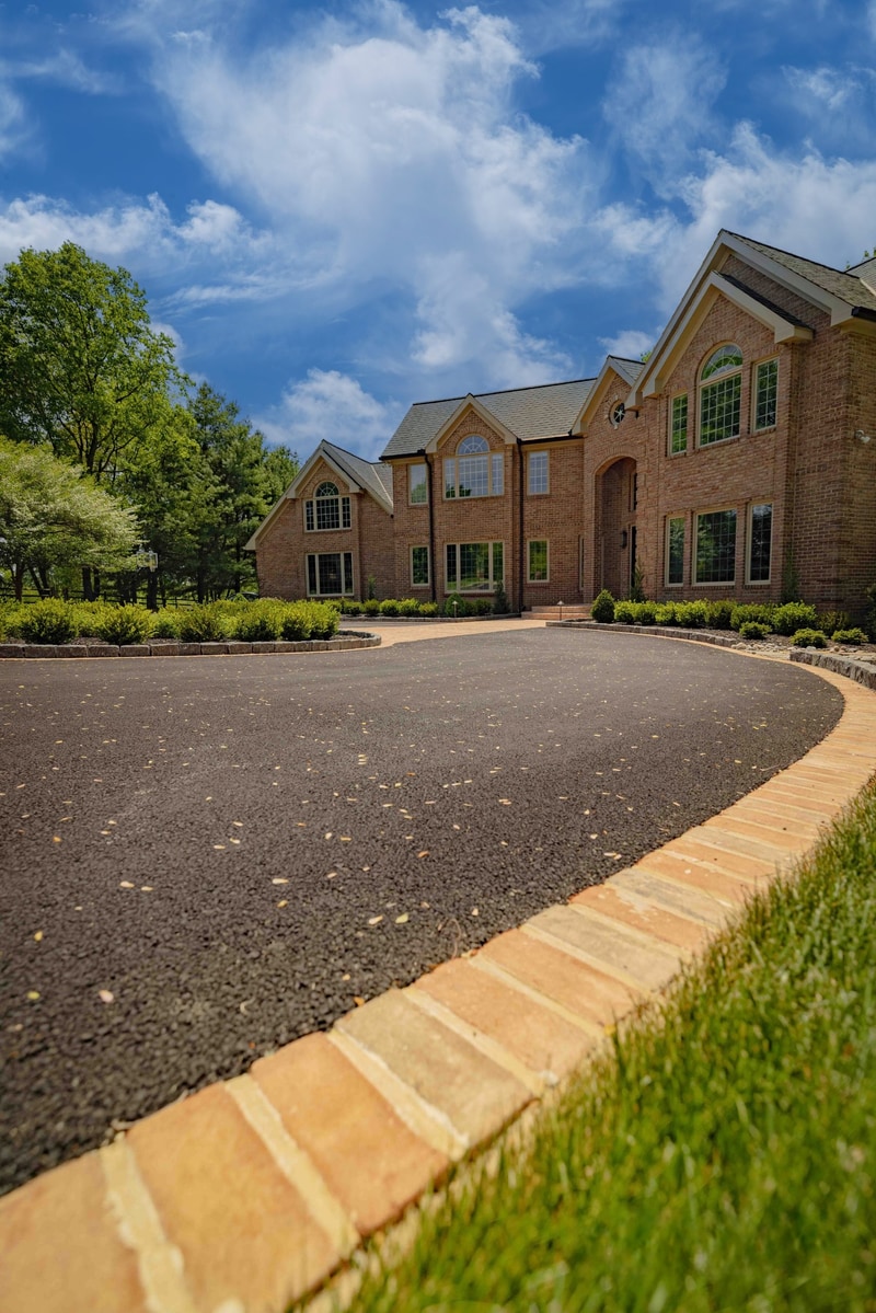 Portrait detail of the Hockessin paver walkway curving past the boxwood front beds: a sun-lit brick paver path with curved cobble edging leads toward the entry courtyard, with the brick facade visible in the background