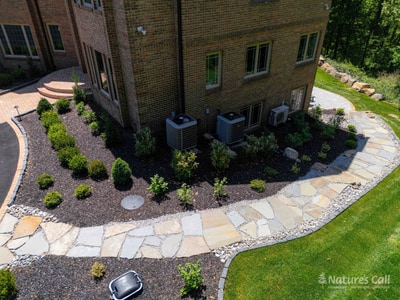 Aerial side view of the Hockessin home: a curved flagstone walkway with brick paver edging threads past the HVAC equipment area, with a river-rock drainage strip running along the foundation and mounded boxwoods softening the utility zone