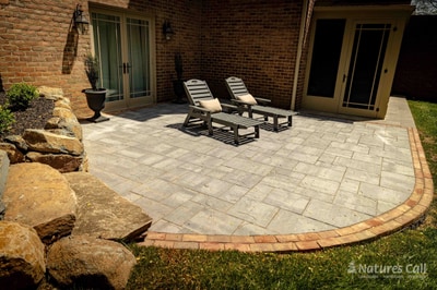Eye-level view of the Hockessin back patio: a tumbled gray paver patio with a brick band edge holds two dark wood chaise lounges against the brick exterior wall, with French doors at the back of the home and rough boulder borders defining the patio's curved edge