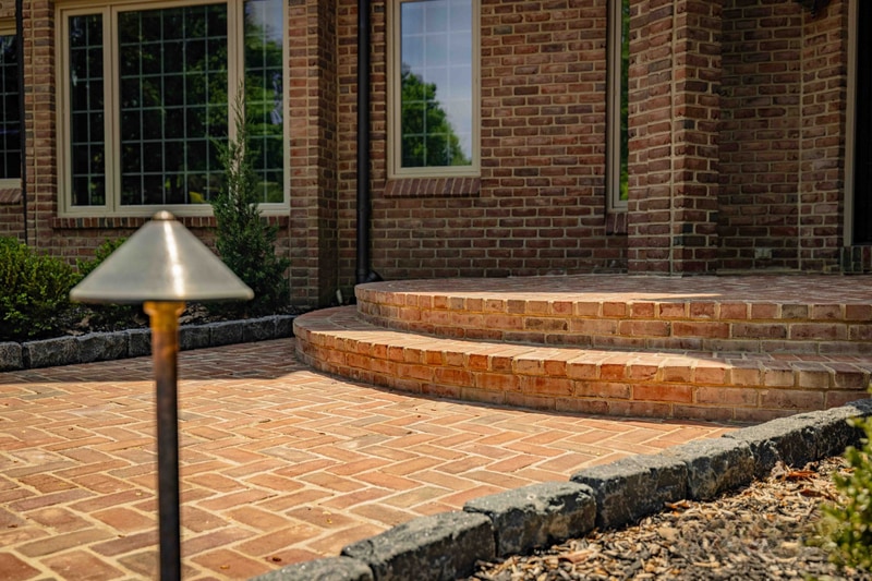 Side-yard view of the Hockessin flagstone path leading toward the woodland edge: irregular flagstones set into a mulched bed are bordered by a low brick edge, with mature trees forming a green backdrop and a clear blue sky overhead