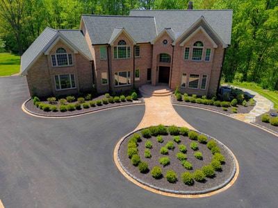Hockessin Delaware estate after Nature's Call rebuild: brick paver walkway and entry, fresh charcoal asphalt driveway with brick borders, brick-edged boxwood-planted oval centerpiece, and refreshed mulched foundation beds with manicured boxwoods.
