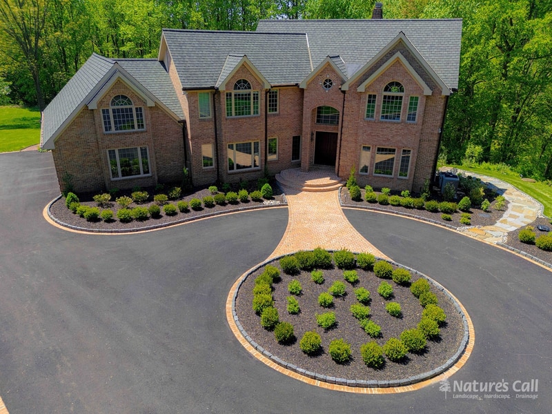 After — Hockessin estate brick home with rebuilt brick paver walkway and entry, fresh charcoal asphalt driveway with brick borders, brick-edged boxwood-planted oval centerpiece, and refreshed mulched foundation beds with manicured boxwoods — Nature's Call Landscaping