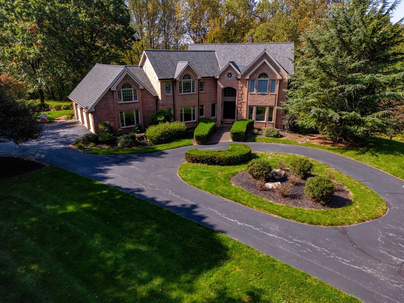 Before — Hockessin estate brick home with original asphalt circular driveway, an oval center bed of loose stones and patchy plantings, and basic foundation shrubs along the front of the house — Nature's Call Landscaping