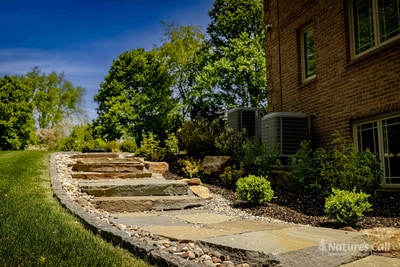 Ground-level view of the Hockessin front entry detail: a brick paver step landing in the foreground, an arched front door with sidelights, a small river-rock drainage strip at the foundation, and mounded boxwoods in the bed alongside