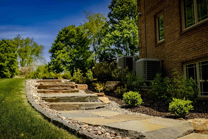 Ground-level view of the Hockessin front entry detail: a brick paver step landing in the foreground, an arched front door with sidelights, a small river-rock drainage strip at the foundation, and mounded boxwoods in the bed alongside